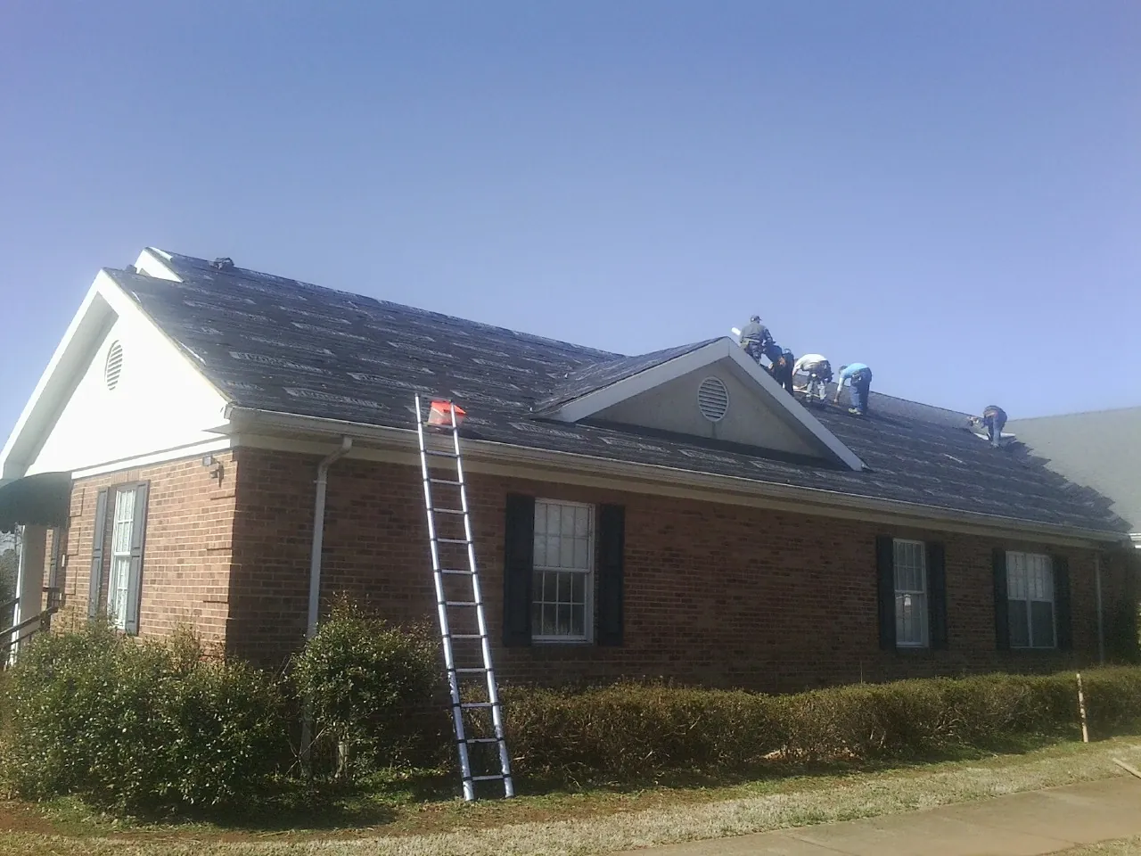 Crew tearing shingles off a brick ranch roof with a ladder set — Metal Roof Repair work by our crew in Polk