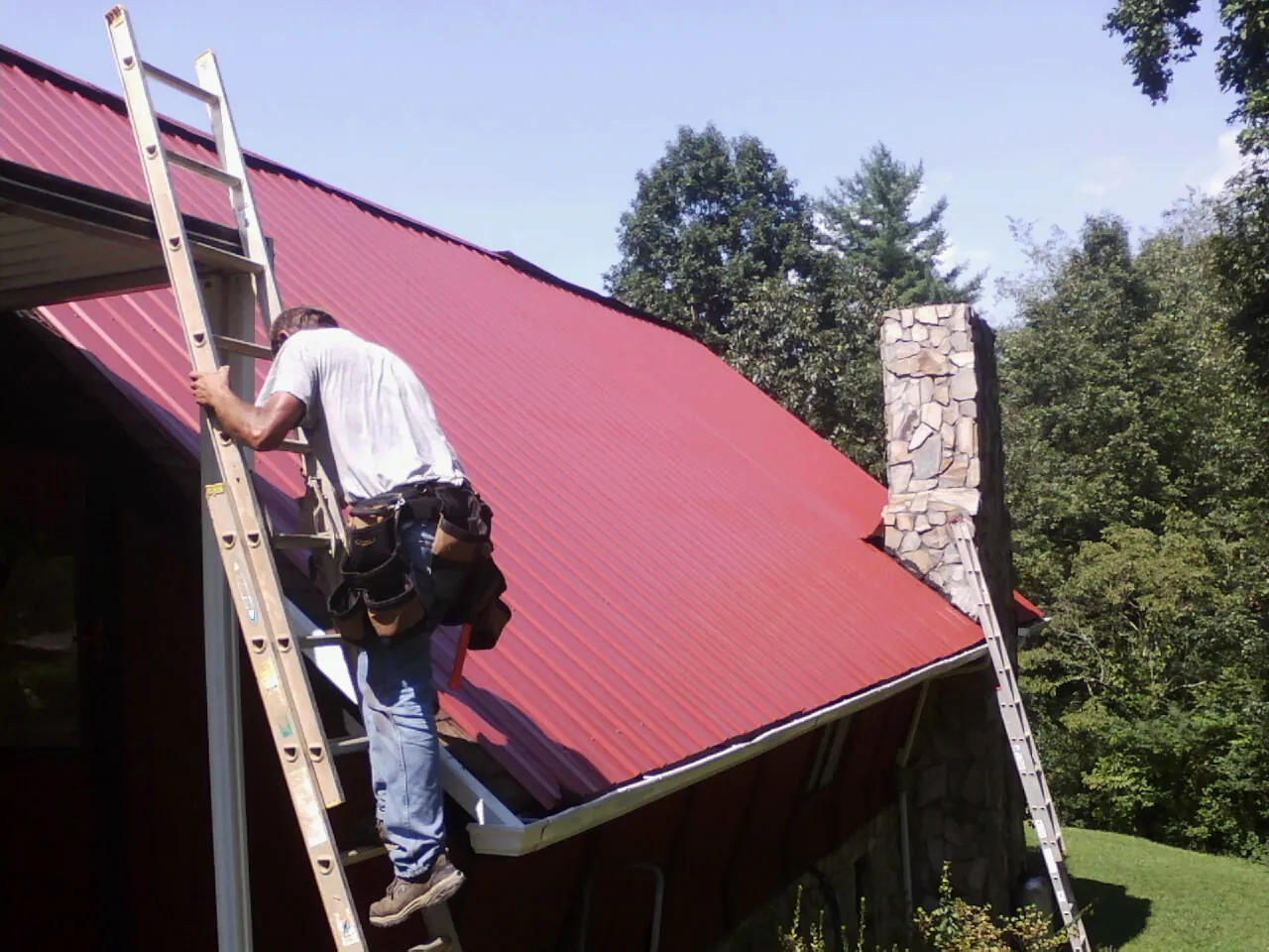 Roofer on a ladder beside a red metal roof and stone chimney — Soffit & Fascia Repair by Roofing Brothers in Polk